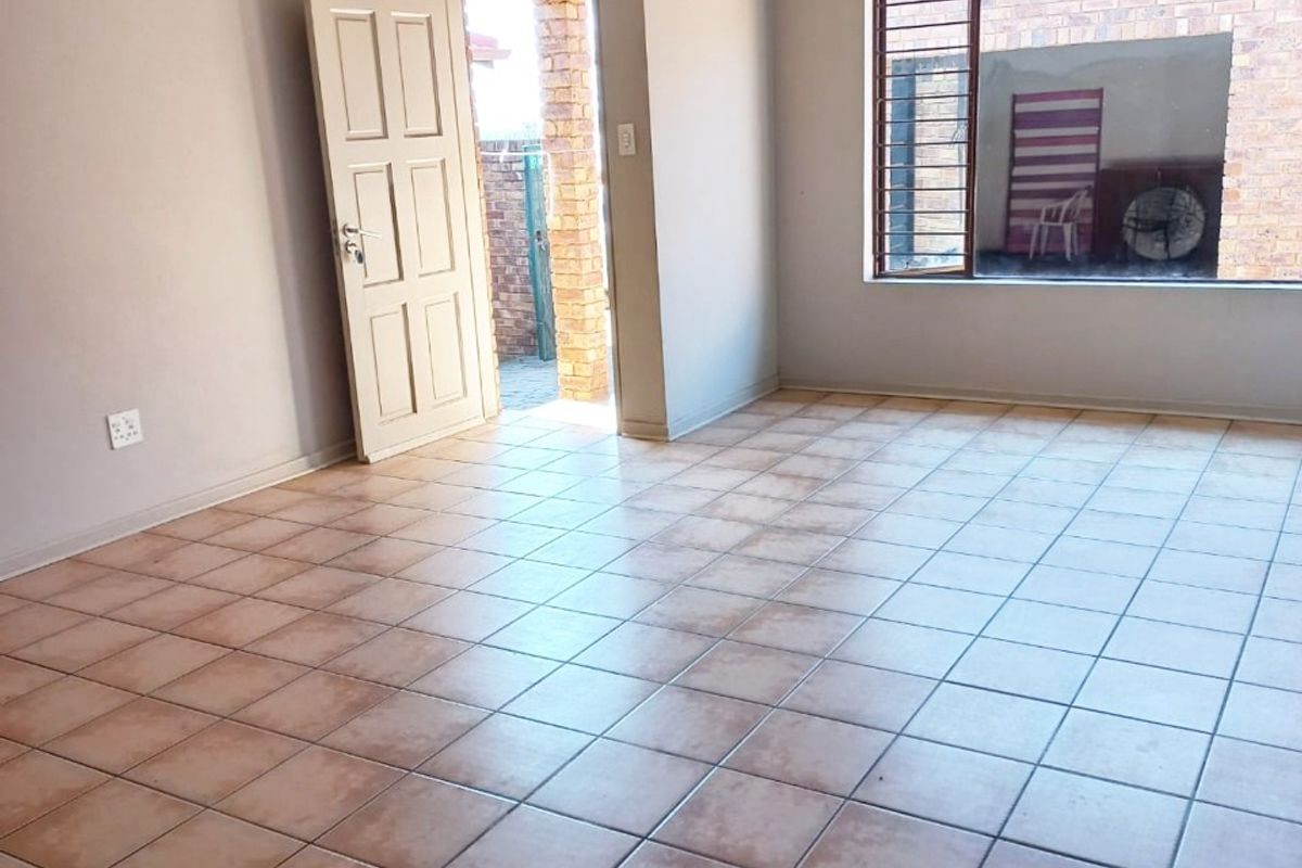 A spacious, empty living area featuring neutral wall tones, tiled flooring, and a large window with security bars, providing access to an outdoor space.