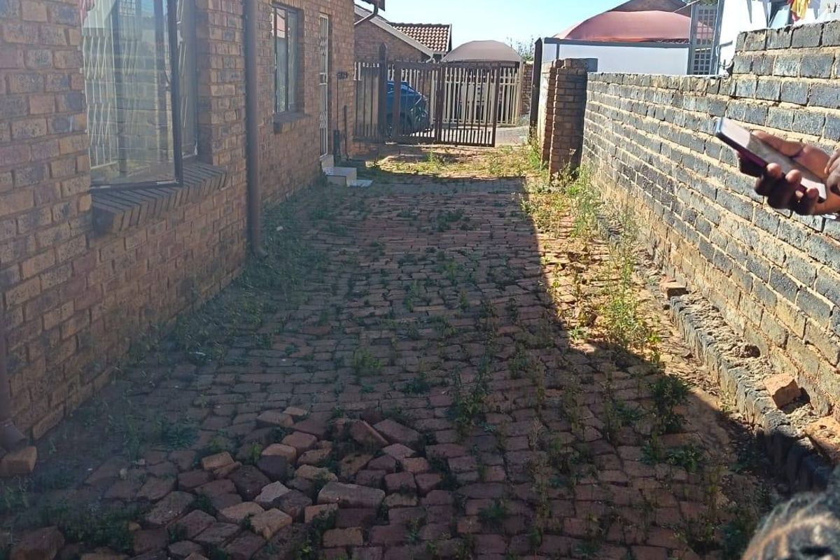 A narrow side or rear exterior passage featuring brick paving, a boundary wall, and a security gate, showing signs of wear and maintenance needs.