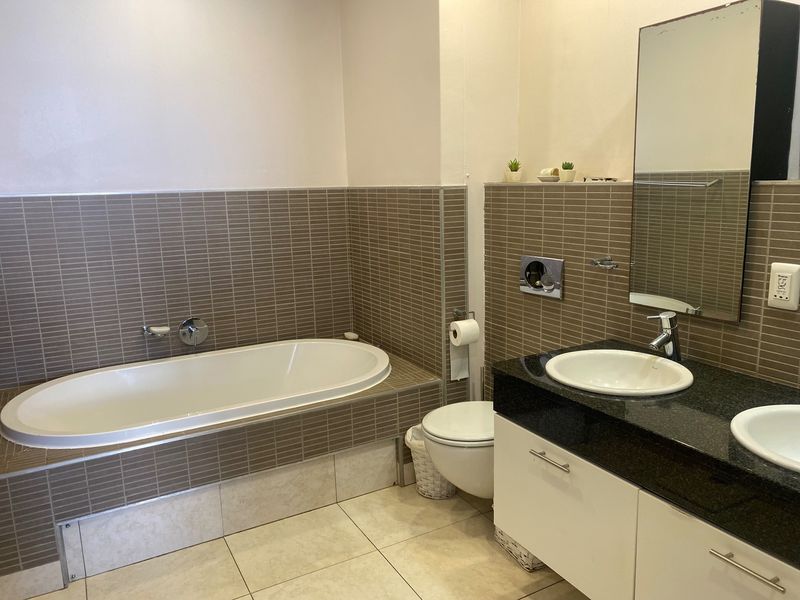 A functional bathroom featuring a built-in bathtub, wall-mounted toilet, and a double vanity with granite countertops and tiled wall finishes.