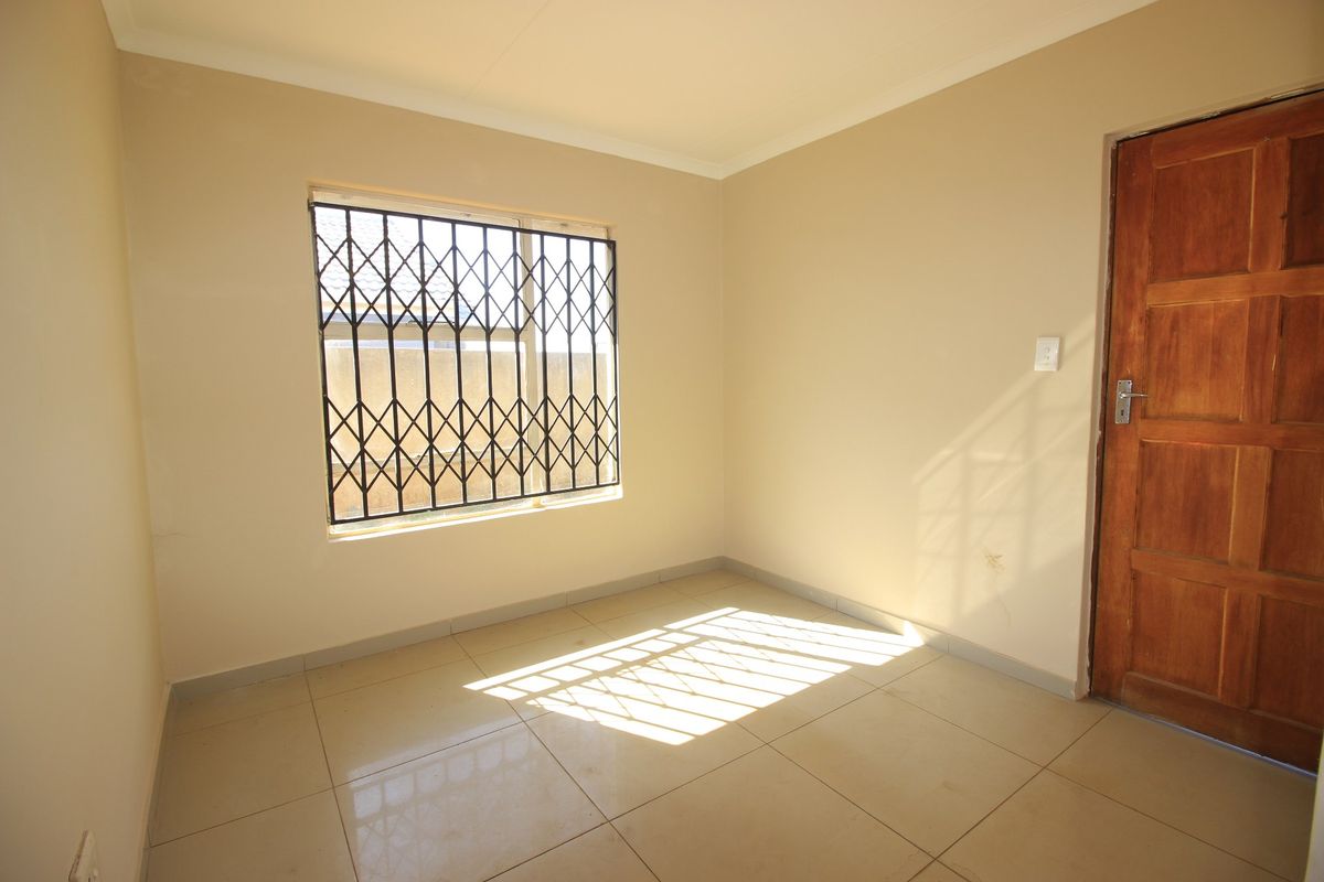 A clean, empty bedroom featuring neutral tiled flooring, a wooden door, and a window fitted with a security gate.