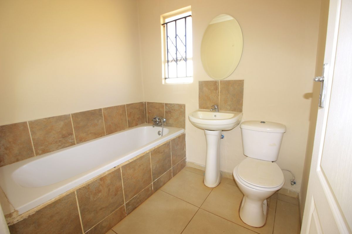 A functional bathroom featuring a bathtub, pedestal basin, and toilet with neutral tiled finishes and a window with security bars.