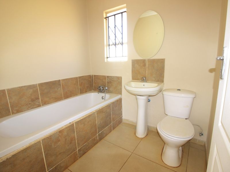 A functional bathroom featuring a bathtub, pedestal basin, and toilet with neutral tiled finishes and a window with security bars.