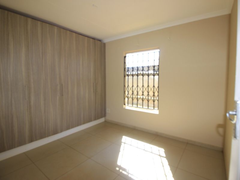 A clean, empty bedroom featuring neutral tiled flooring, built-in wooden cupboards, and a window fitted with a security gate.