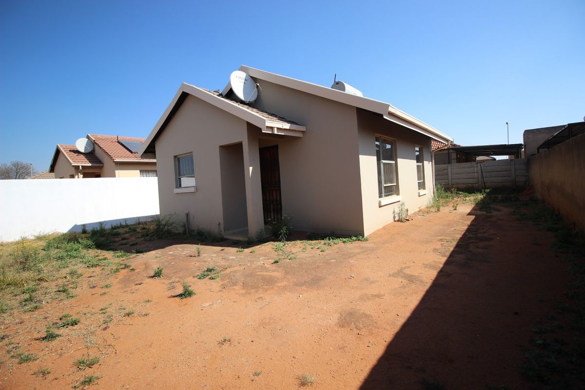 Rear view of a detached house featuring a tiled roof, satellite dish, solar geyser, and a large, unlandscaped yard enclosed by a precast concrete wall.