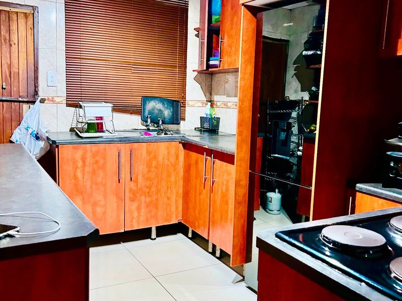 A functional kitchen featuring warm-toned laminate cabinetry, dark countertops, ceramic tiled flooring, and an integrated electric hob with an extractor fan.