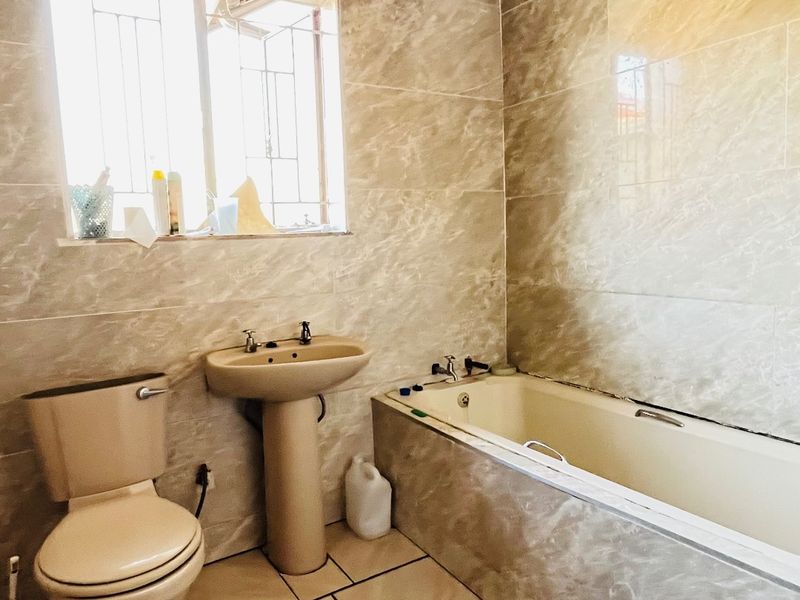 A functional bathroom featuring full-height wall tiling, a pedestal basin, a bathtub, and a toilet, with security bars installed on the window.