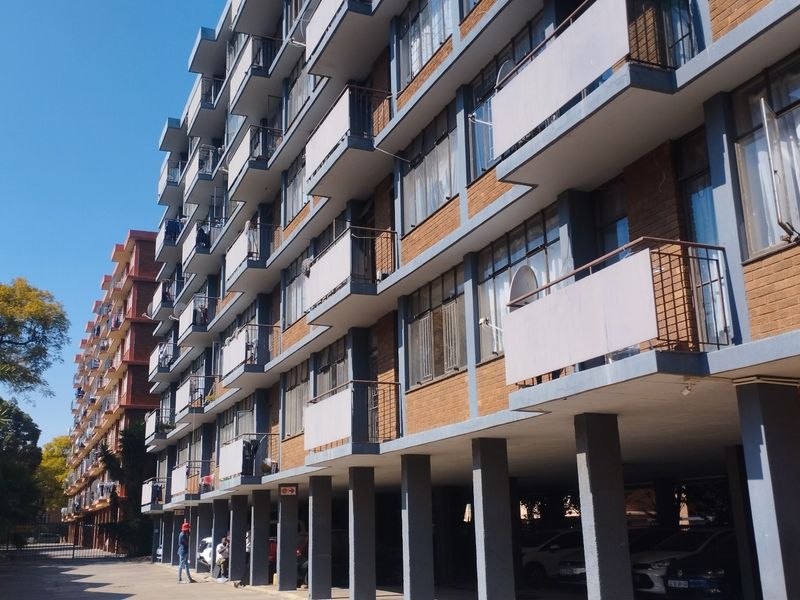 Exterior view of a multi-story apartment building featuring brick facade, private balconies with metal railings, and a ground-level covered parking area.