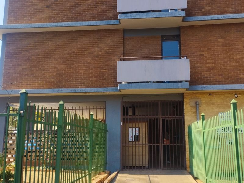 Front elevation of a multi-story brick apartment building featuring balconies, secure gated entrance, and perimeter fencing with security wire.