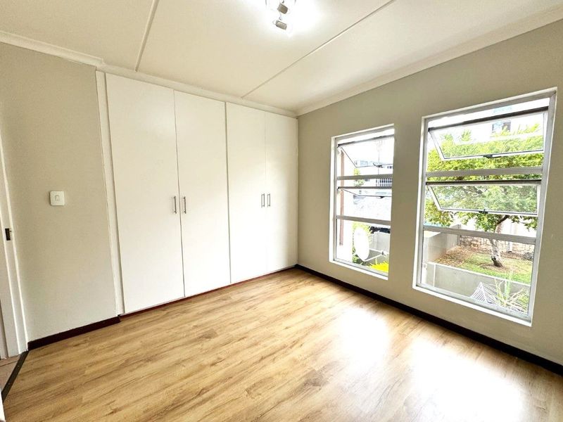 A bright, empty bedroom featuring light wood-look laminate flooring, ample built-in cupboard space, and large aluminium-framed windows overlooking the garden.