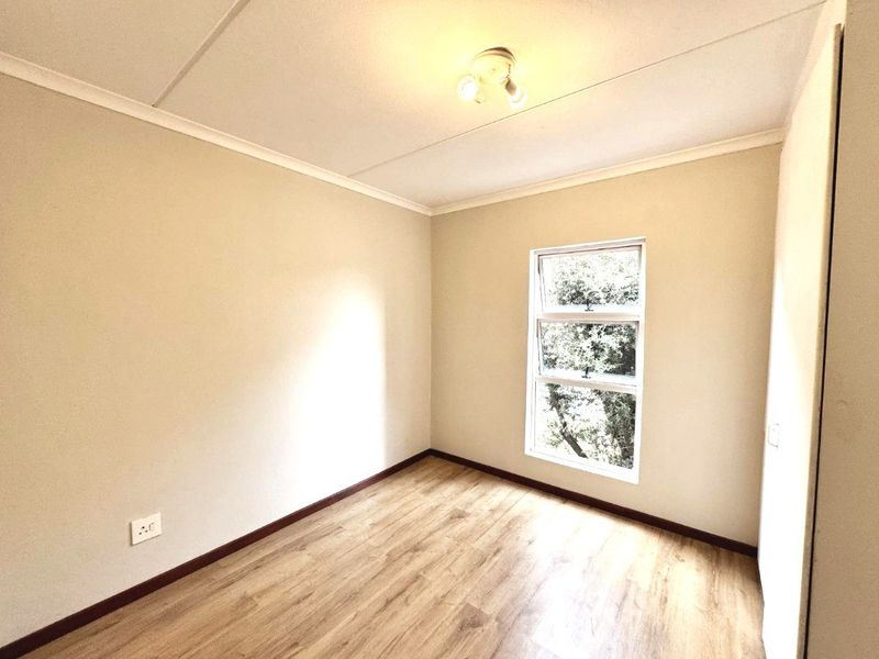 A bright, empty bedroom featuring modern laminate flooring, a built-in cupboard, and a window providing natural light.