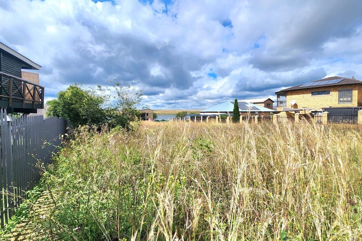 A view of a vacant residential plot featuring overgrown grass and surrounding perimeter fencing.