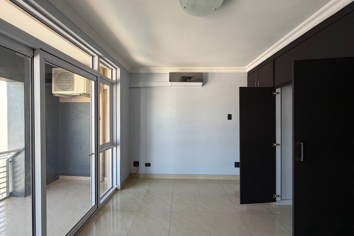 A bright, empty bedroom featuring tiled floors, built-in dark cabinetry, a split-system air conditioner, and a large sliding glass door leading to a balcony.