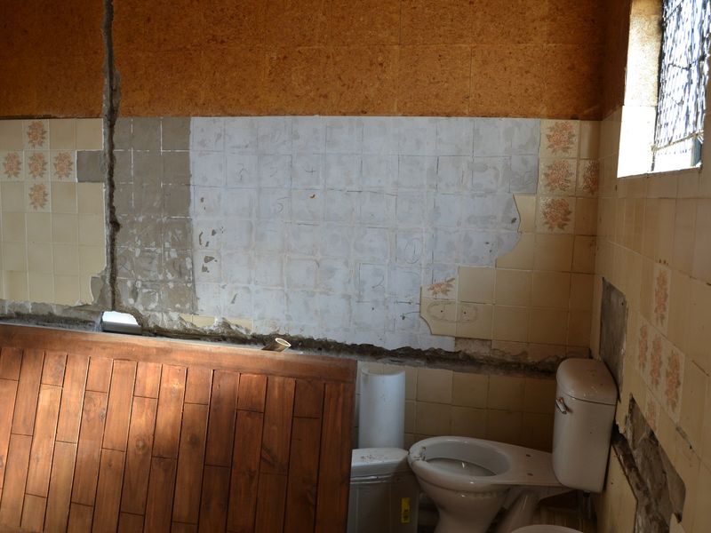A bathroom in need of significant renovation, featuring damaged wall tiles, exposed plaster, and a basic toilet fixture.