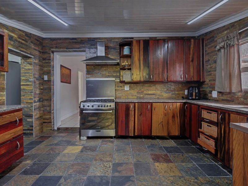 A rustic kitchen featuring stone-clad walls, slate flooring, wooden cabinetry, granite countertops, and a stainless steel gas stove with an extractor fan.
