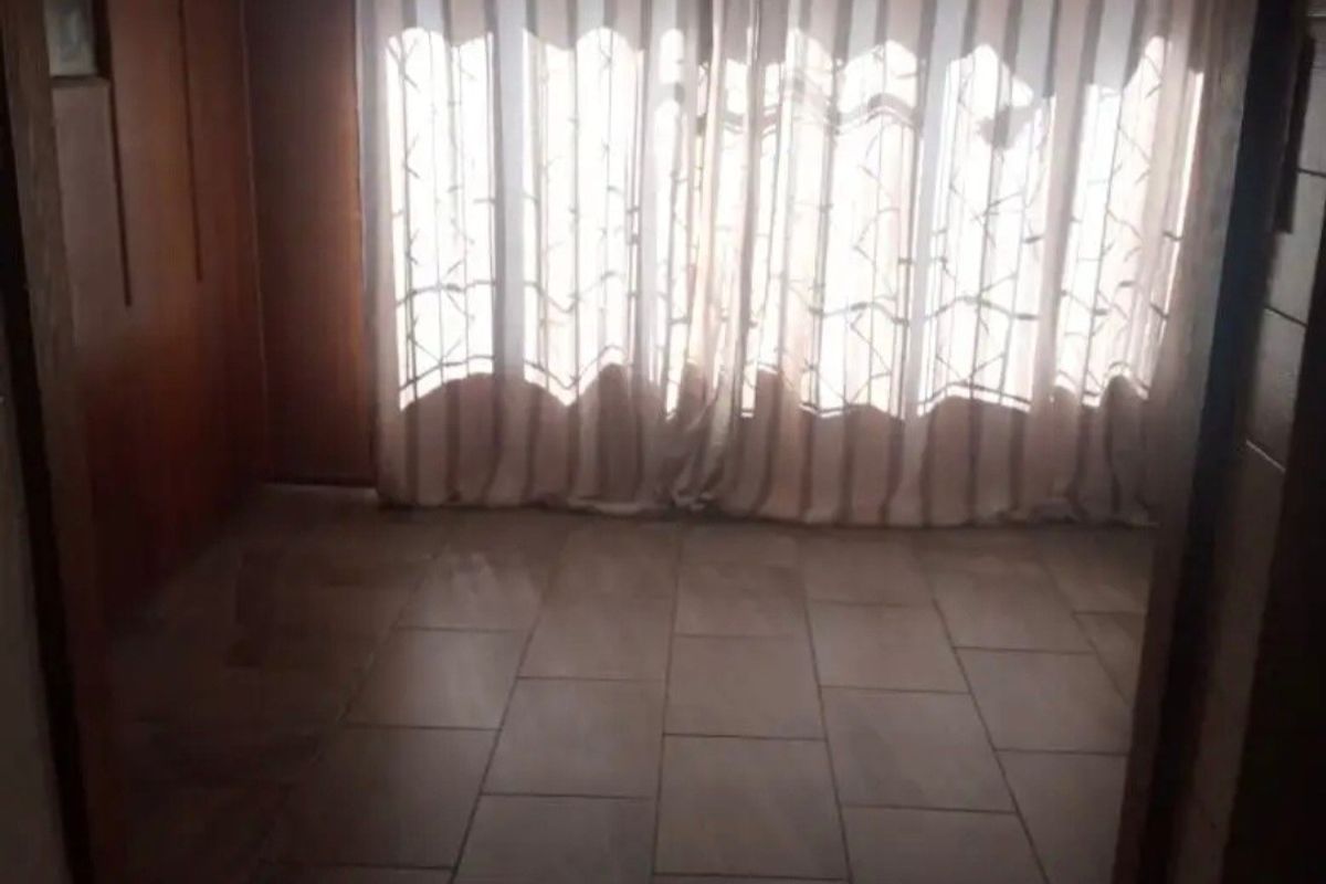 An empty living area featuring tiled flooring and wood-paneled walls, illuminated by natural light through large window curtains.