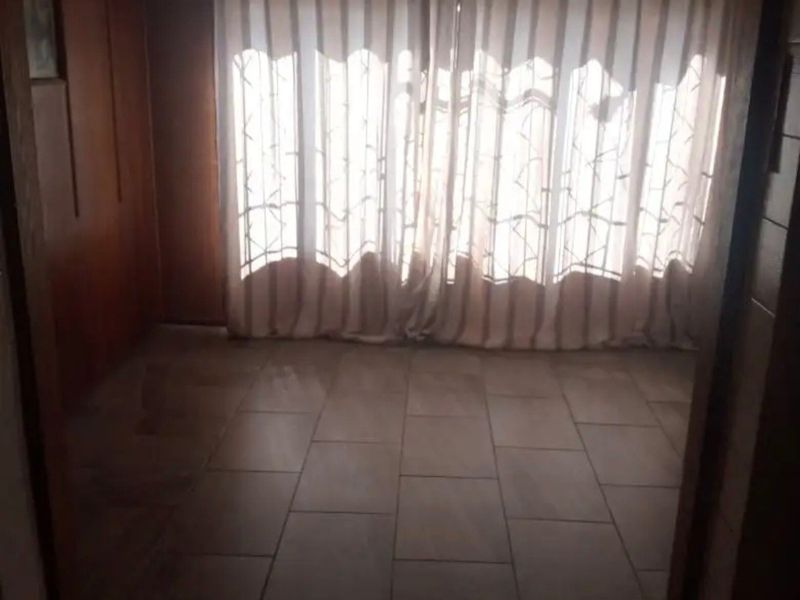 An empty living area featuring tiled flooring and wood-paneled walls, illuminated by natural light through large window curtains.