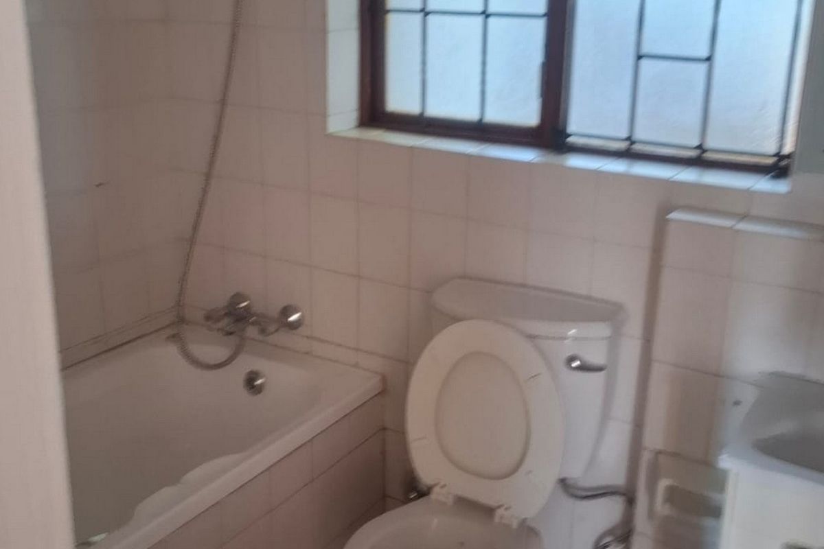 A dated bathroom featuring a built-in bathtub, toilet, and vanity basin, with tiled walls and a window fitted with security bars.