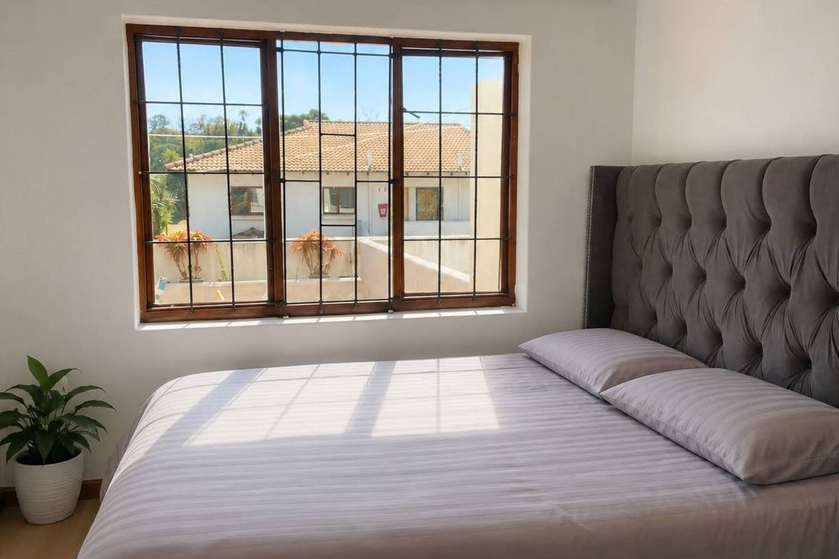 A bright bedroom featuring laminate flooring and a large window with security bars, offering a view of the surrounding townhouse complex.