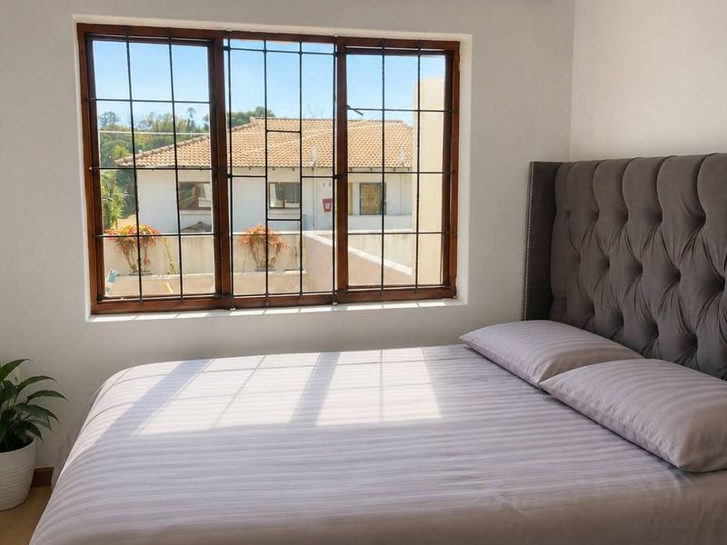 A bright bedroom featuring laminate flooring and a large window with security bars, offering a view of the surrounding townhouse complex.