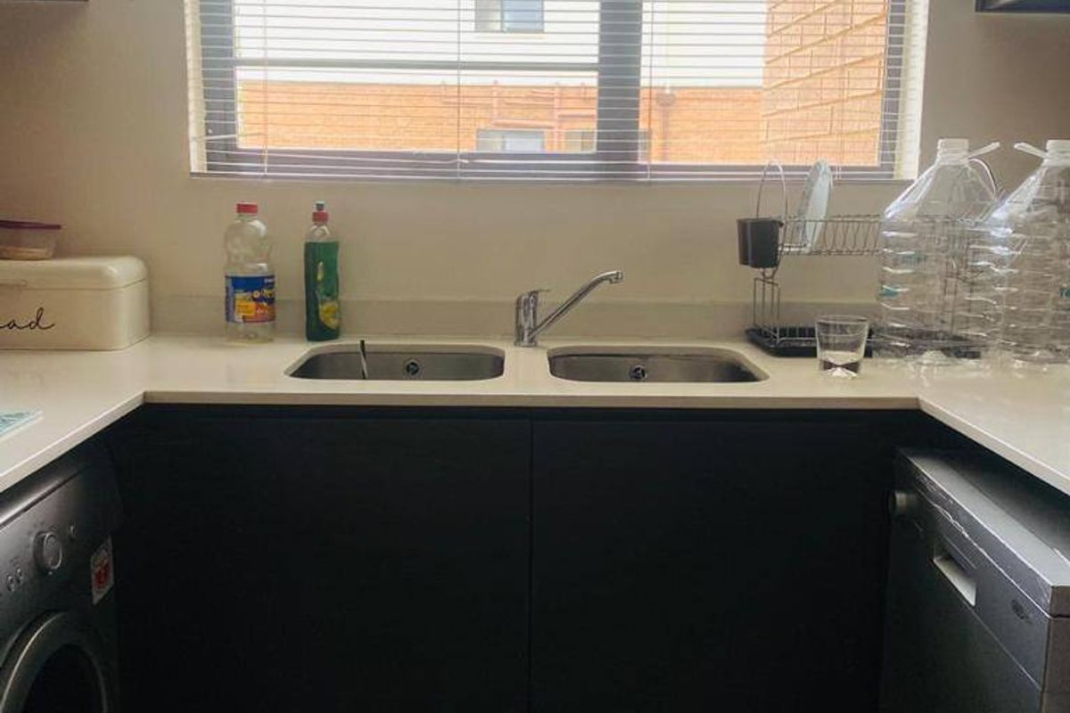 A functional kitchen area featuring dark cabinetry, stone countertops, a double bowl sink, and tiled flooring, illuminated by a window with venetian blinds.