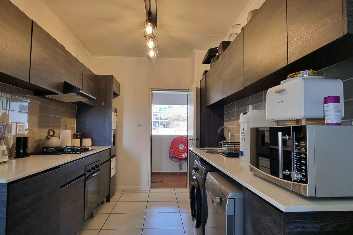 Modern galley-style kitchen featuring dark wood-grain cabinetry, stone countertops, gas hob, built-in oven, and tiled flooring with direct access to a balcony.