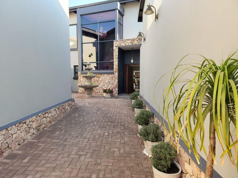 A well-maintained paved entrance walkway featuring stone cladding, a decorative water fountain, and large glass windows leading to the main entrance.