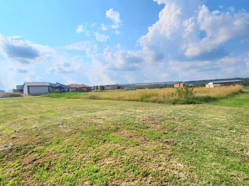 A wide-angle view of a vacant residential plot featuring an open grass lawn with surrounding neighborhood houses in the distance.