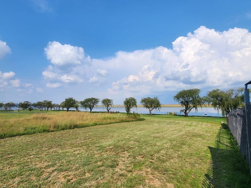 A scenic view of a vacant residential plot featuring open grassland, mature trees, and a distant water body.