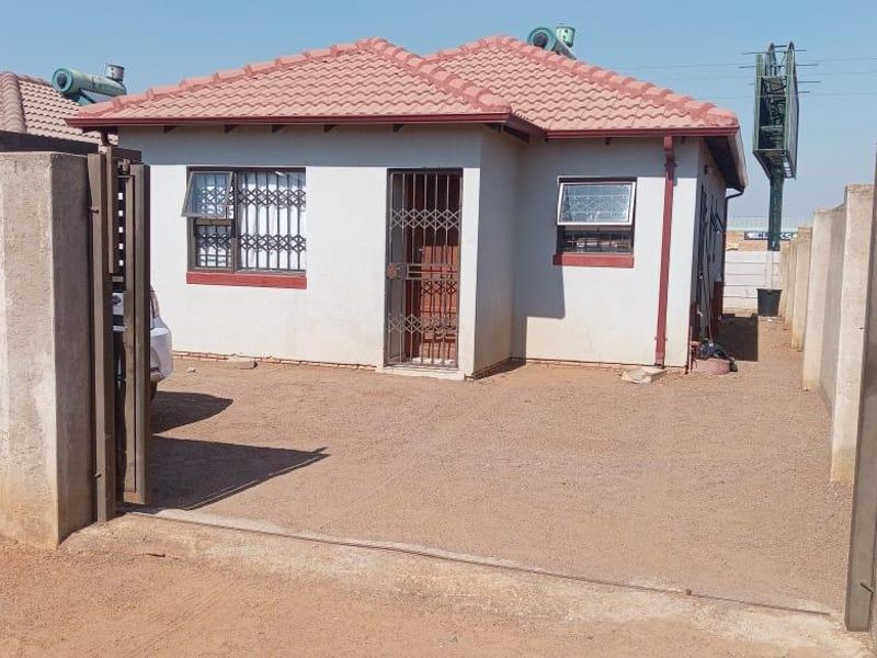 Front view of a single-story house featuring a tiled roof, security gates, burglar bars, and a perimeter wall with an unpaved driveway.
