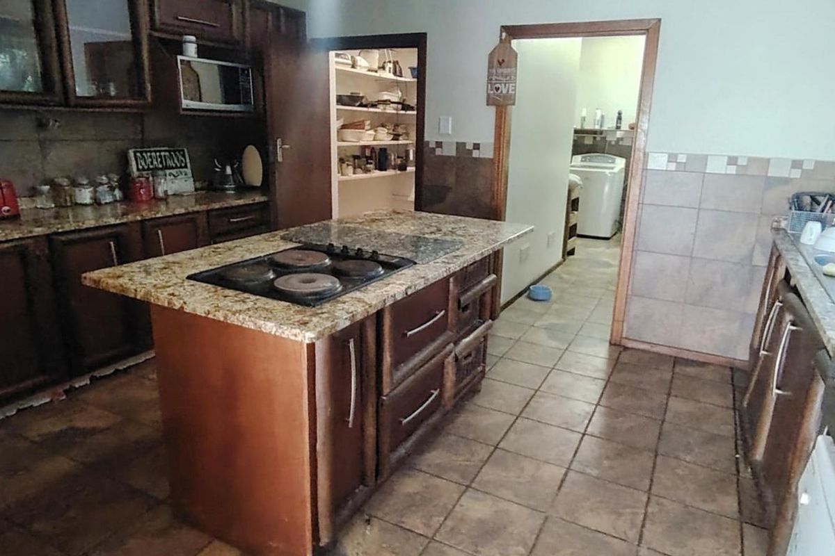 A functional kitchen featuring granite countertops, a central island with a built-in hob, ample wooden cabinetry, and an adjacent pantry area.