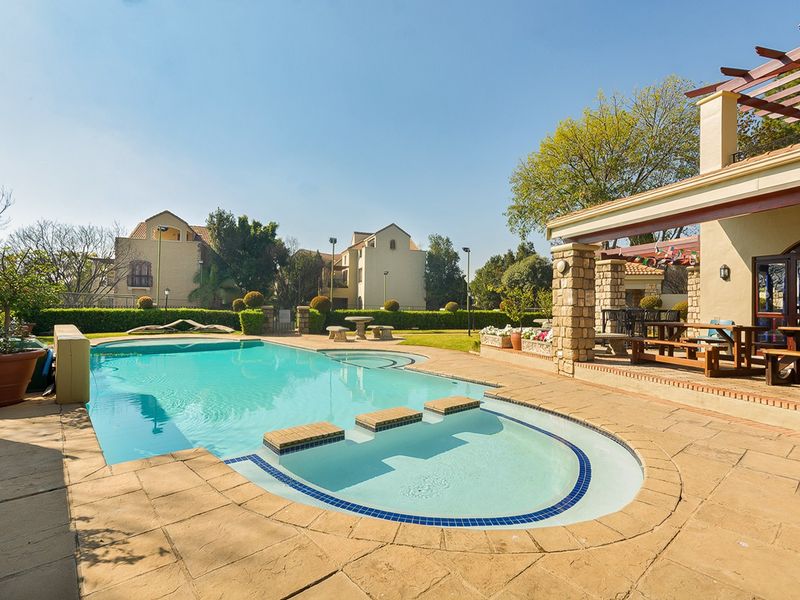 A well-maintained outdoor pool area featuring a paved patio, stone-clad pergola structure, and landscaped garden surroundings.