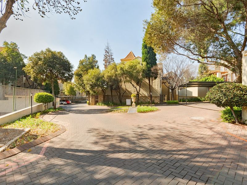 A well-maintained paved driveway and entrance area of an apartment complex, featuring mature trees, landscaped garden beds, and covered parking bays.