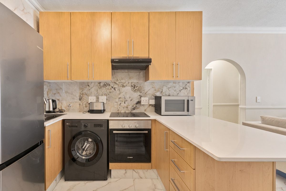 Modern kitchen featuring light wood cabinetry, stone countertops, marble-look backsplash, built-in oven, electric hob, and an integrated washing machine, leading into an arched hallway.