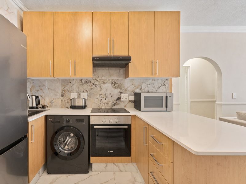 Modern kitchen featuring light wood cabinetry, stone countertops, marble-look backsplash, built-in oven, electric hob, and an integrated washing machine, leading into an arched hallway.