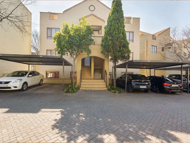 Front elevation of an apartment complex featuring a paved driveway, covered carport parking bays, and a central staircase leading to the building entrance.