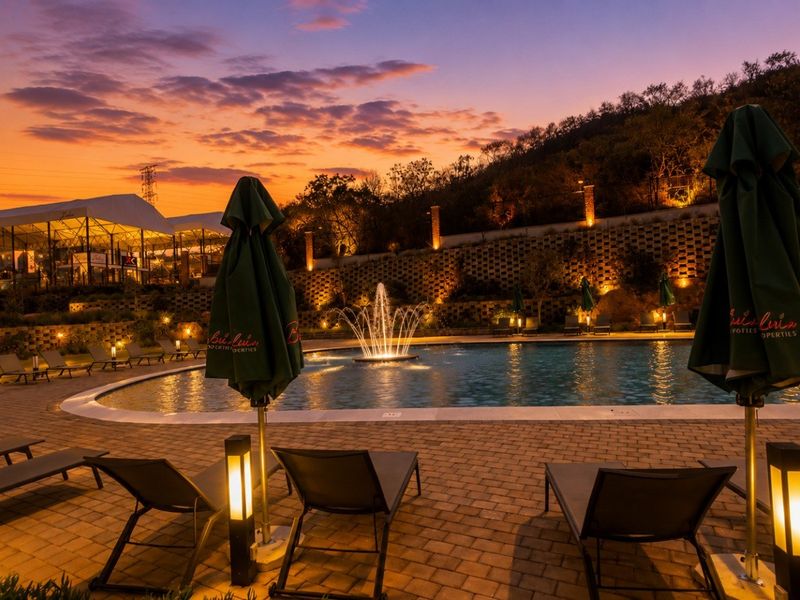 A beautifully landscaped communal pool area featuring a central water fountain, paved decking, and ambient evening lighting set against a scenic hillside backdrop.