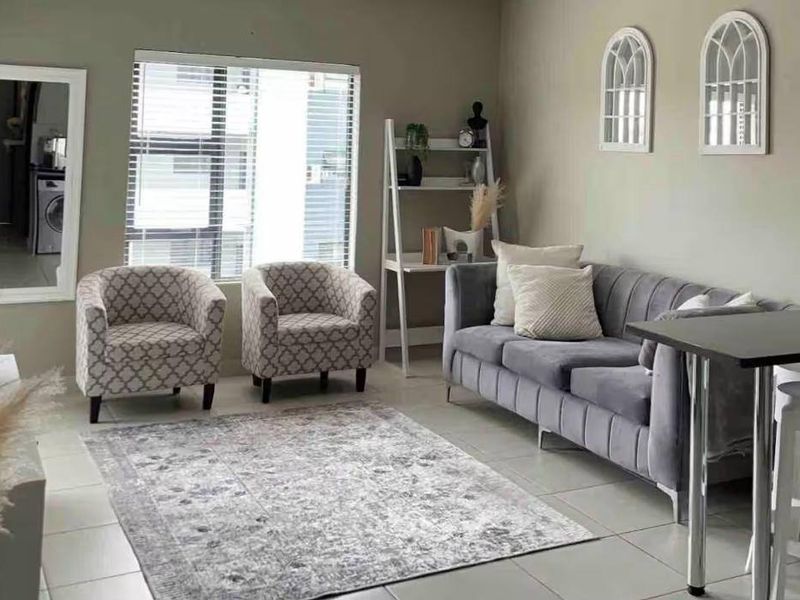 A well-maintained living room featuring neutral wall tones, tiled flooring, and large windows with venetian blinds, offering a bright and comfortable living space.