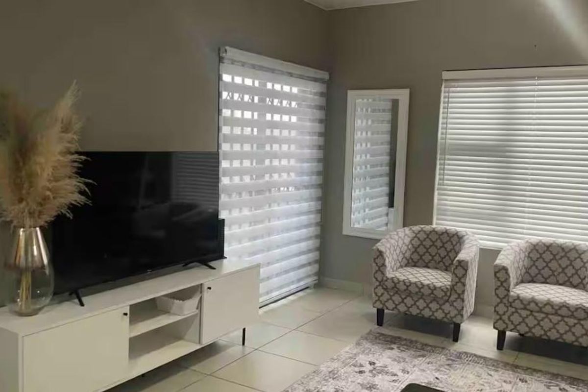 A well-maintained living room featuring neutral wall tones, tiled flooring, and dual window treatments including venetian and roller blinds.