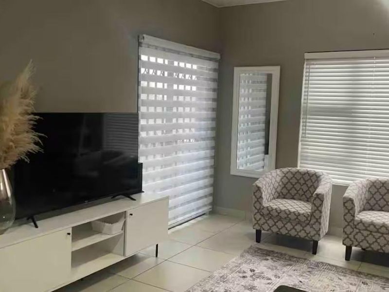 A well-maintained living room featuring neutral wall tones, tiled flooring, and dual window treatments including venetian and roller blinds.