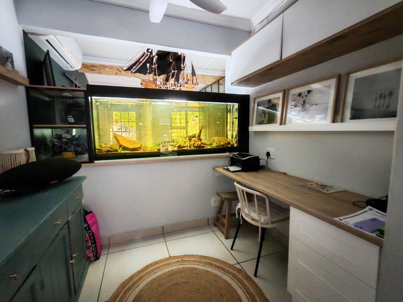 A functional home office featuring built-in desk space, overhead cabinetry, wall-mounted shelving, and a ceiling fan, finished with tiled flooring.