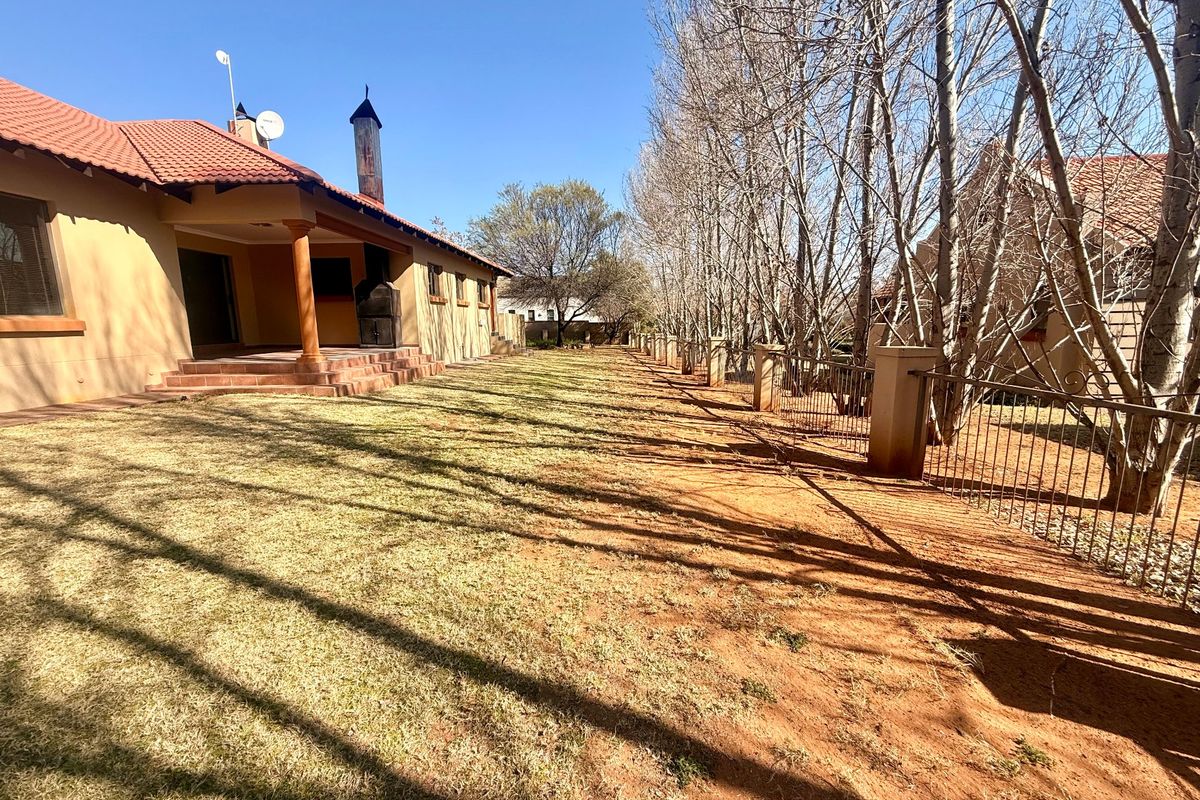 Rear view of the property featuring a covered patio with a built-in braai, overlooking a spacious garden with perimeter metal fencing.