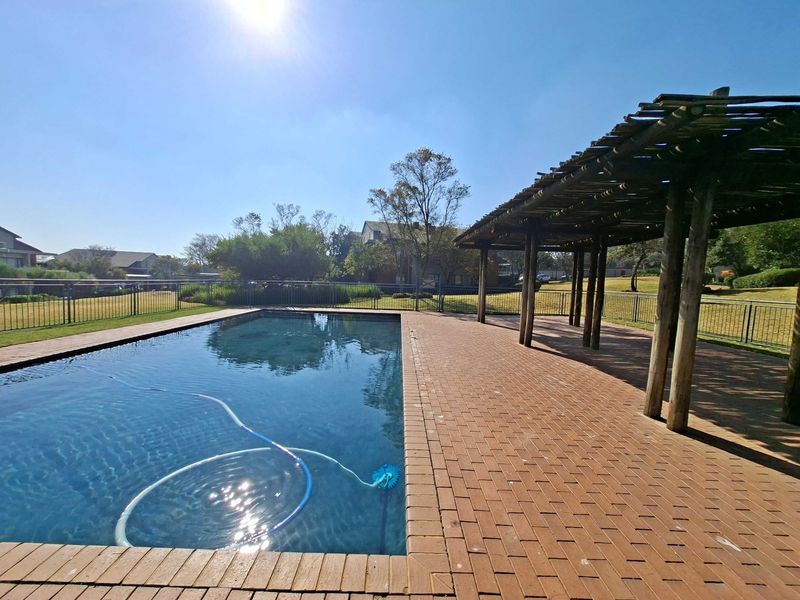 Communal swimming pool area featuring brick paving, a timber pergola for shade, and secure metal perimeter fencing in a well-maintained outdoor setting.