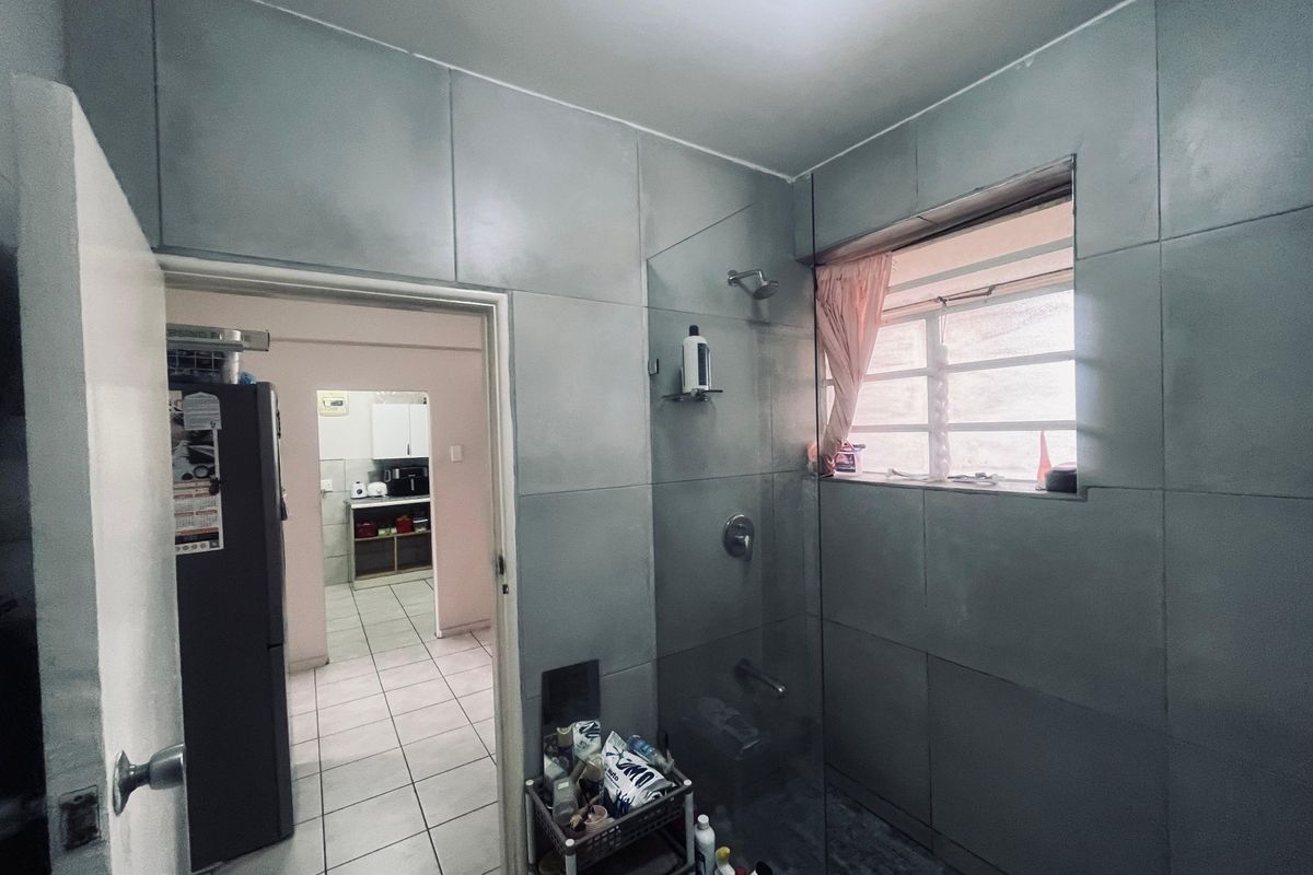 A bathroom featuring full-height grey wall tiles, a glass shower screen, and a louvered window, with a view into the adjacent kitchen area.