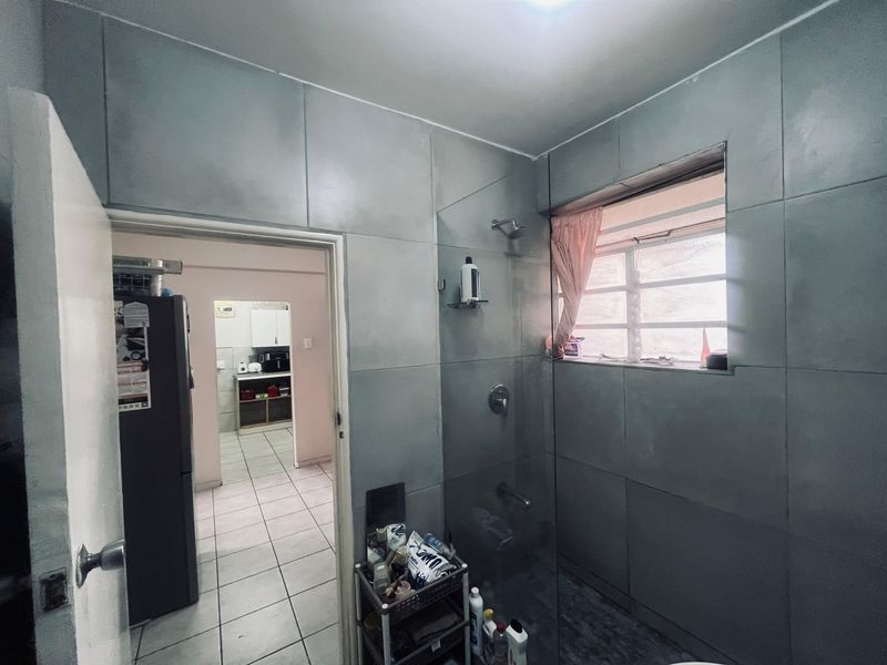 A bathroom featuring full-height grey wall tiles, a glass shower screen, and a louvered window, with a view into the adjacent kitchen area.