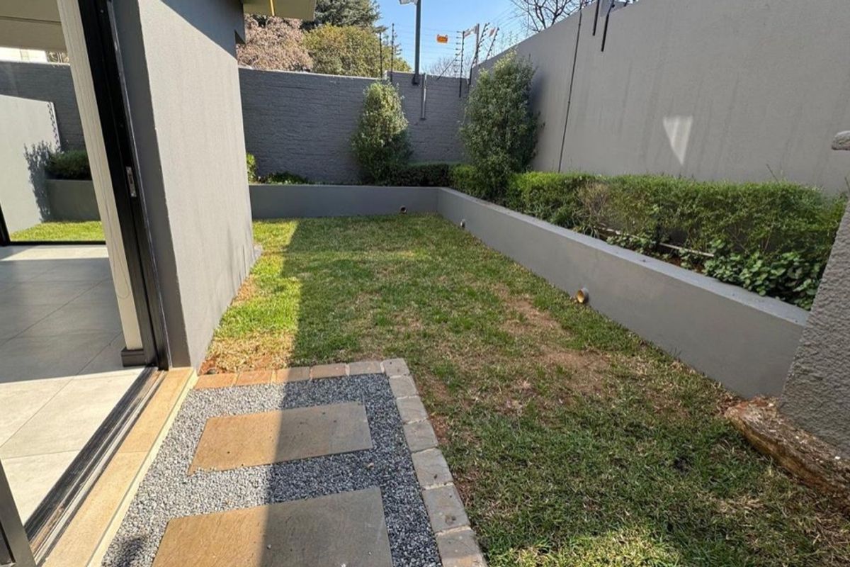 A well-maintained rear garden featuring a paved walkway, manicured lawn, and a solid perimeter wall with integrated electric fencing for added security.