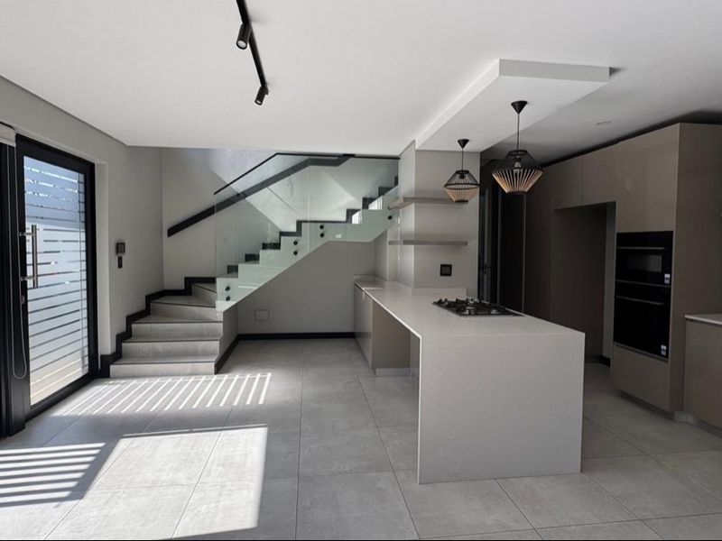 Modern open-plan kitchen featuring a central island with gas hob, built-in oven, sleek cabinetry, and a striking glass balustrade staircase.