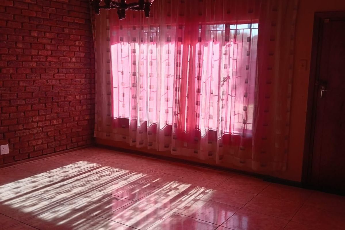 An empty living room featuring a rustic exposed brick wall, timber ceiling, and tiled flooring, illuminated by natural light through large windows.