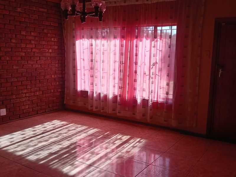 An empty living room featuring a rustic exposed brick wall, timber ceiling, and tiled flooring, illuminated by natural light through large windows.