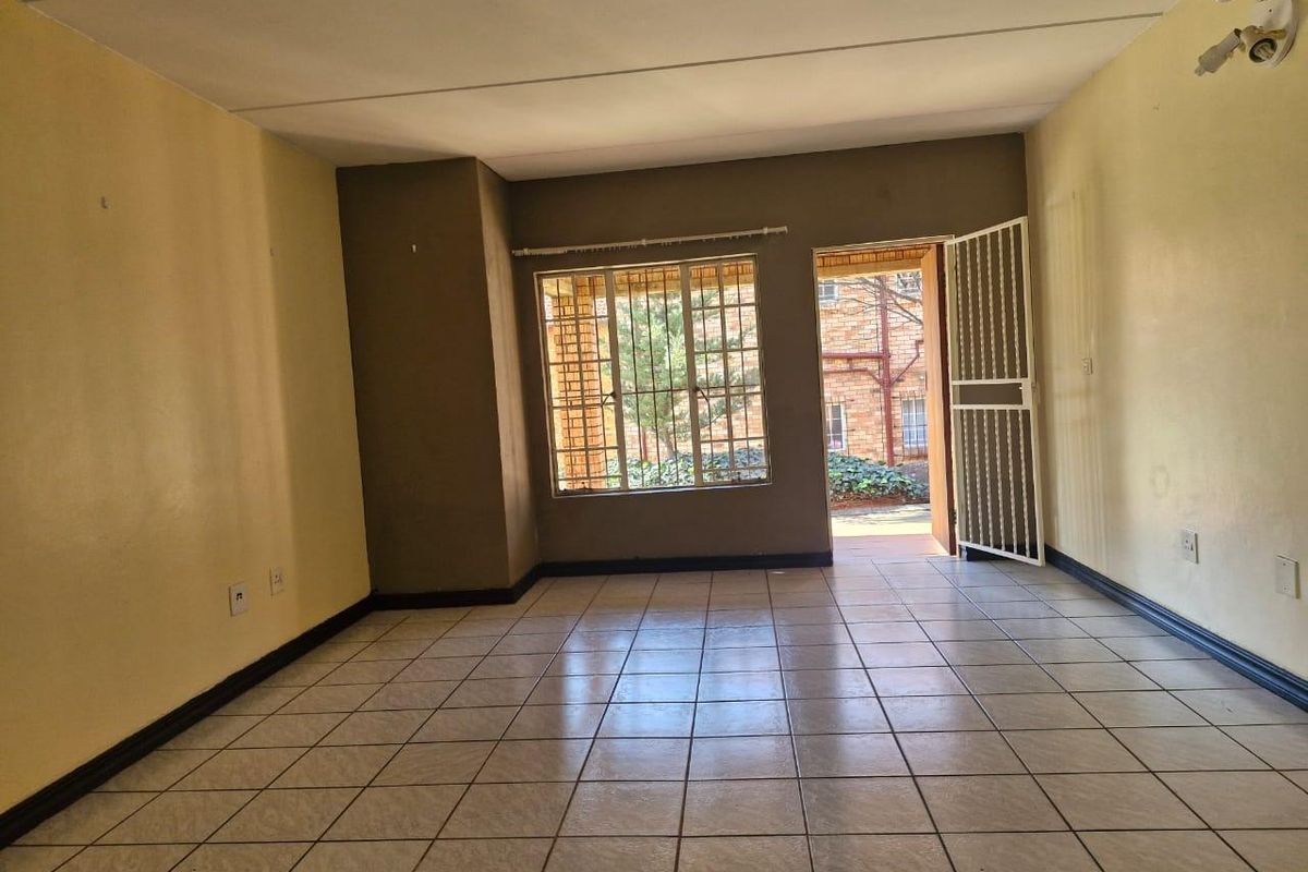 An empty, open-plan living area featuring tiled flooring, security gate access, and large windows with burglar bars, ready for a fresh coat of paint.