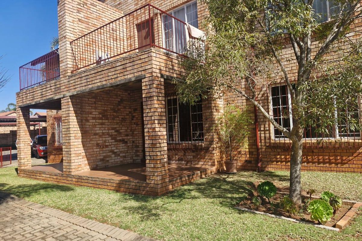 A well-maintained face brick townhouse featuring a covered ground-floor patio, an upper-level balcony with metal railings, and a neat garden area.